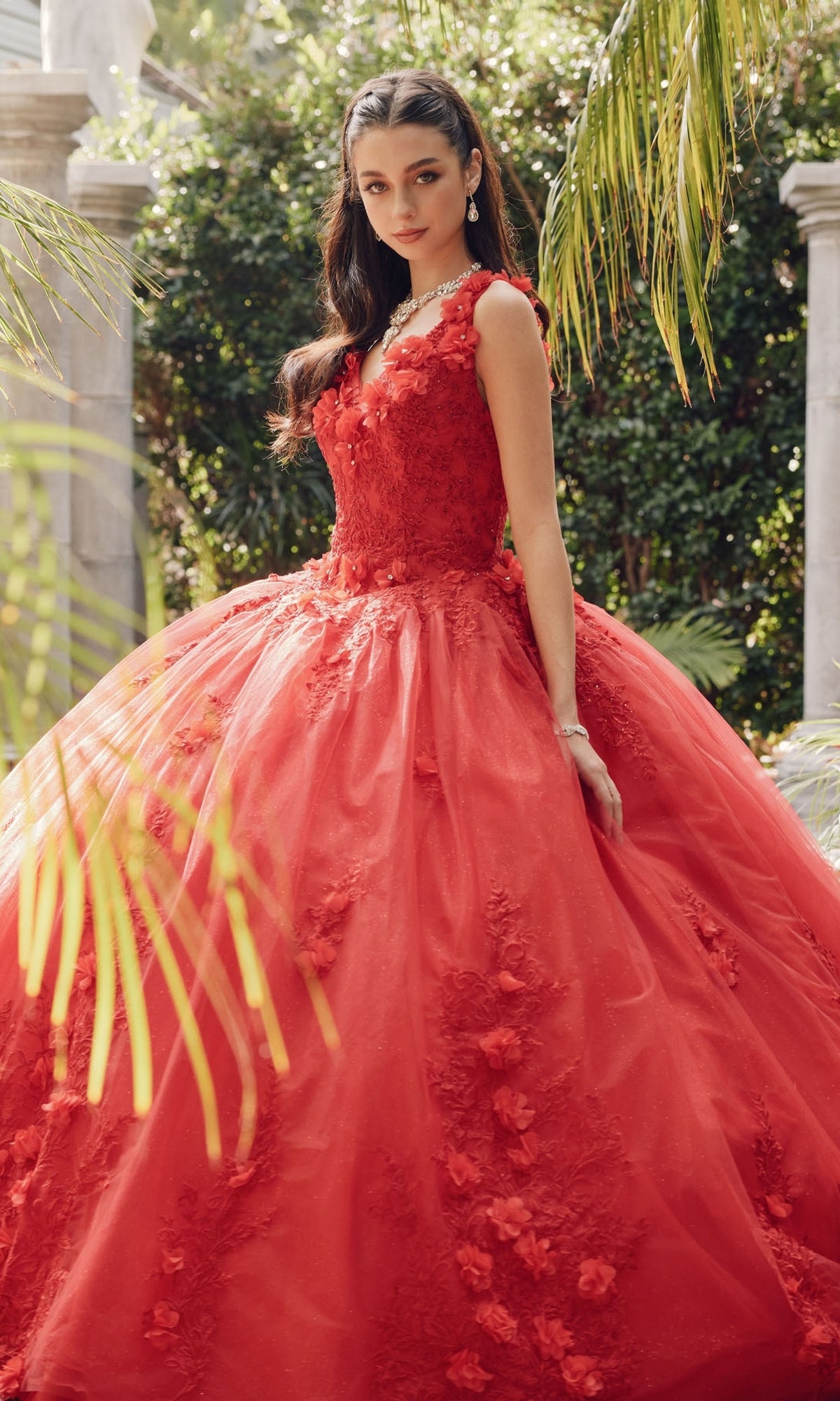 A woman stands outside in the Juliet 1437R Red Ball Gown Quinceañera Dress, featuring floral details, beaded embroidery, and a full skirt, surrounded by lush greenery and stone columns.